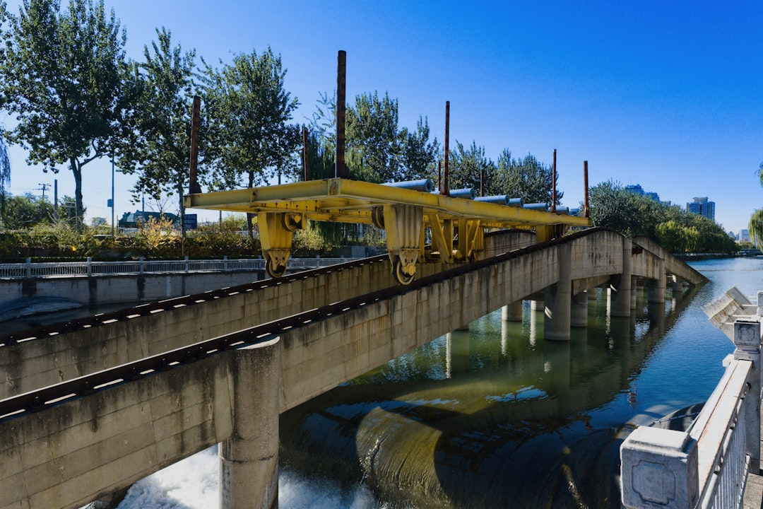 Large yellow train bridge with water below and trees around, showcasing engineering and nature in bright daylight - A striking yellow train bridge spans over a body of water, surrounded by lush green trees. The bright blue sky adds to the scene, highlighting the contrast of nature and engineering.